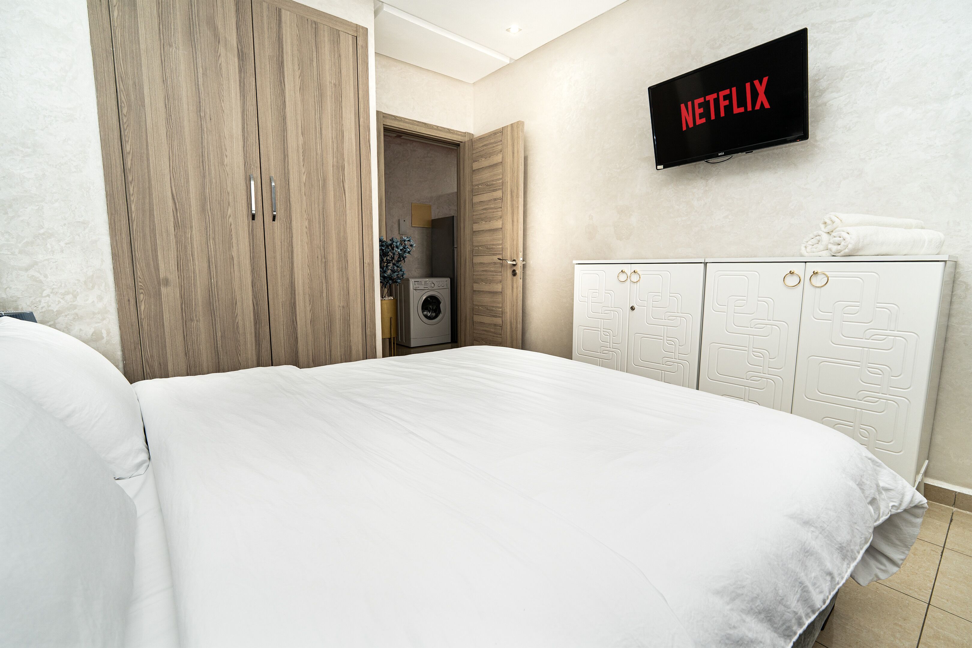 A bedroom with a bed, wardrobe, washing machine, and a TV displaying 'NETFLIX'.