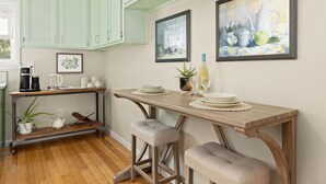 A kitchen with light green cabinets, a wooden table, two barstools, a coffee maker, a kettle, a wine bottle, and two framed paintings. - Cozy, Quiet 2 Bedroom with Hot Tub, Fire Pit, Close to Fayetteville (Winslow)