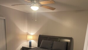 Ein Schlafzimmer mit einem Bett, einem Nachttisch, einer Lampe, einem Deckenventilator und einer Tür.
