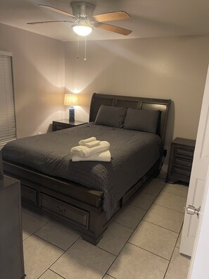 Ein Schlafzimmer mit einem Bett, einem Nachttisch, einer Lampe, einem Deckenventilator und einer Tür.