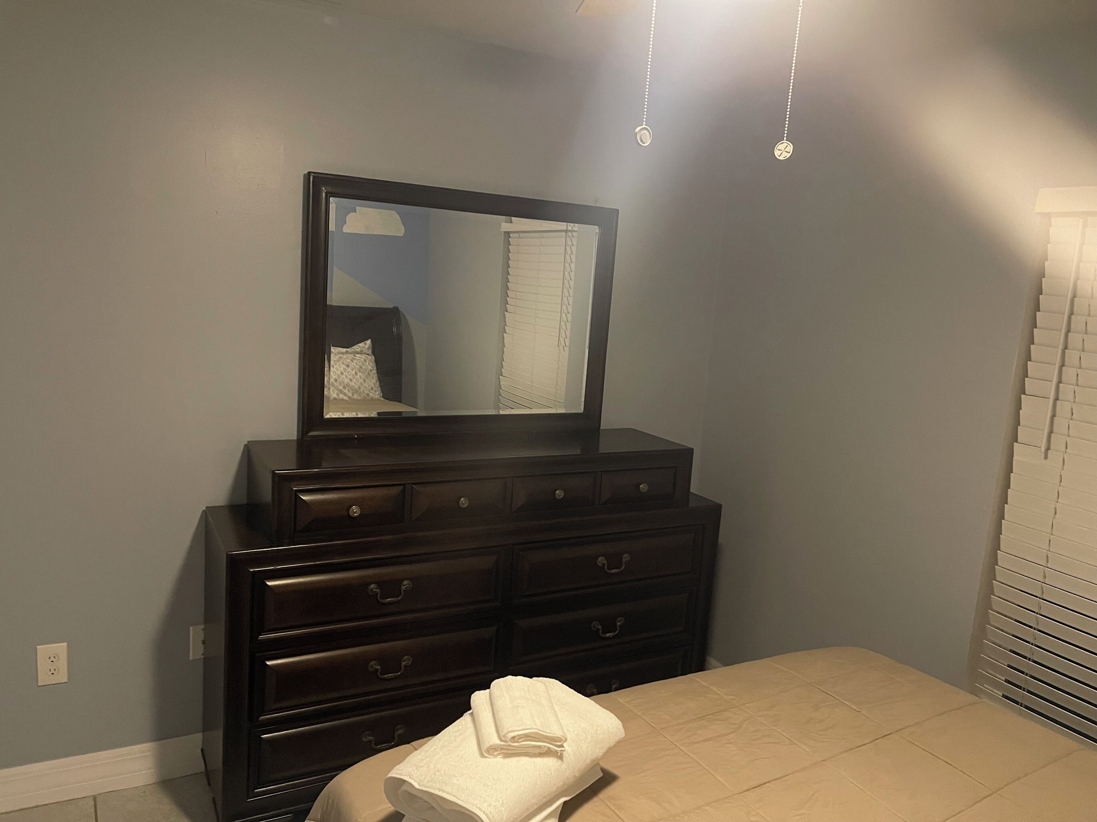 Un dormitorio con un aparador oscuro, un espejo, una cama con colcha color beige y una ventana con persianas.
