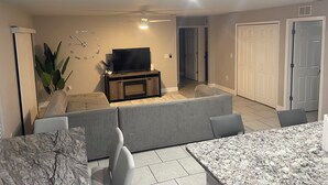 Ein Wohnzimmer mit Fernseher auf einem Holzboden, grauen Sofas, einem Esstisch mit Stühlen und einem Deckenventilator.