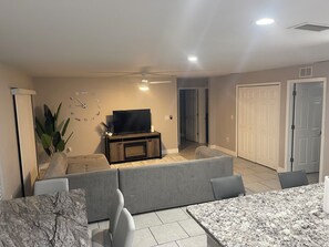 Ein Wohnzimmer mit Fernseher auf einem Holzboden, grauen Sofas, einem Esstisch mit Stühlen und einem Deckenventilator.
