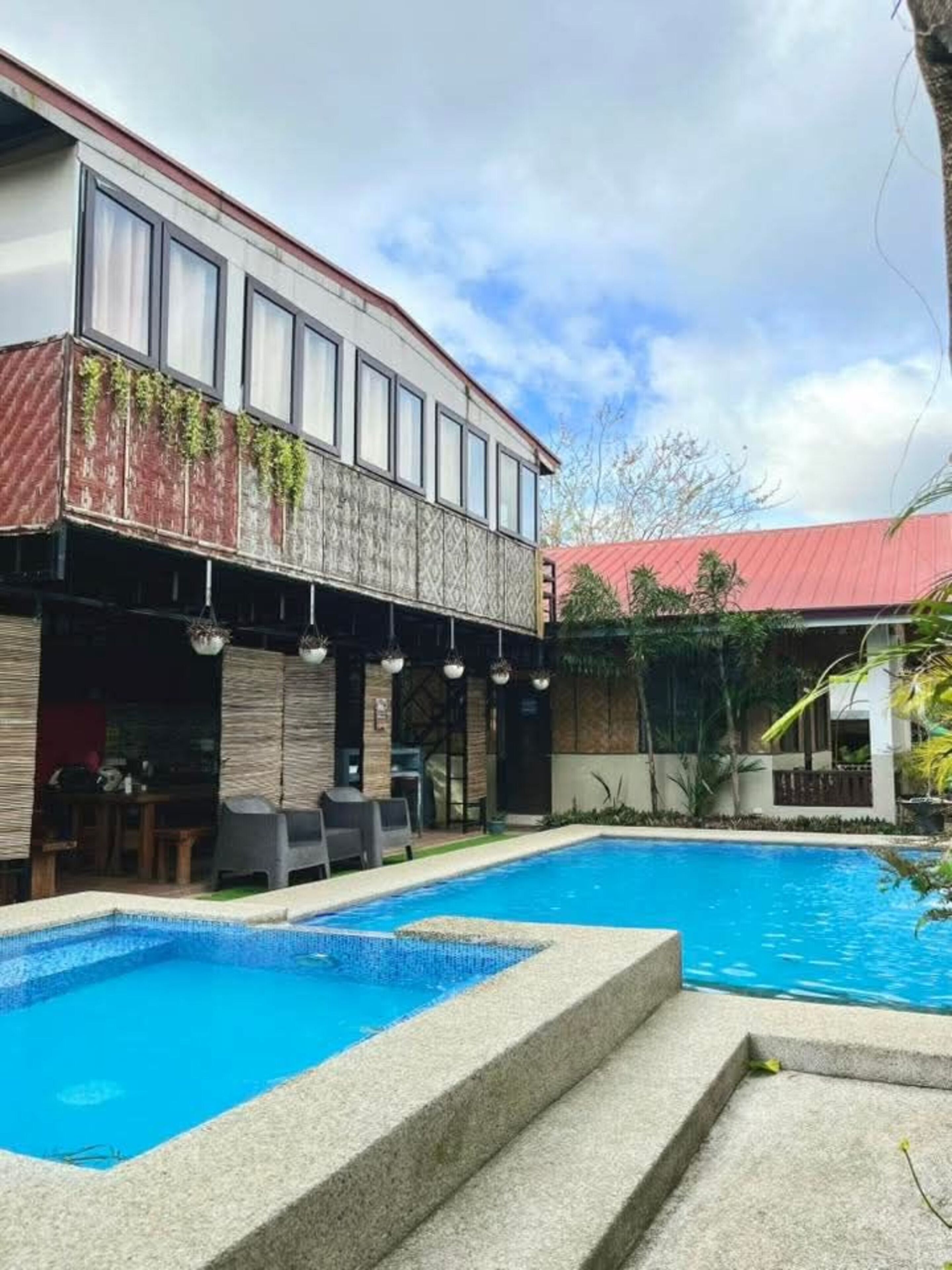 A two-story building with a swimming pool, outdoor seating, and a red-roofed structure in the background.