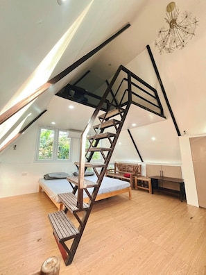 A loft-style room with a wooden staircase, a bed, a desk, and a view of greenery outside.