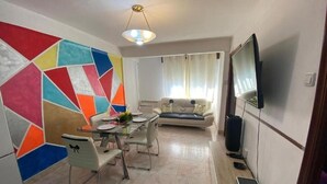 Uma área de jantar com mesa e cadeiras de vidro, um sofá, uma televisão e uma parede com padrão geométrico colorido.