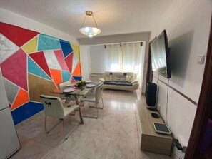 A dining area with a glass table and chairs, a sofa, a television, and a wall with a colorful geometric pattern. - Familia y Grupos Luces Navidad Vigo (Vigo)