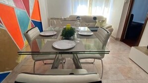 Uma área de refeições com mesa de vidro, cadeiras brancas e uma parede colorida.