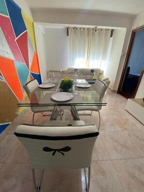 A dining area with a glass-top table, white chairs, and a colorful wall. - Familia y Grupos Luces Navidad Vigo (Vigo)