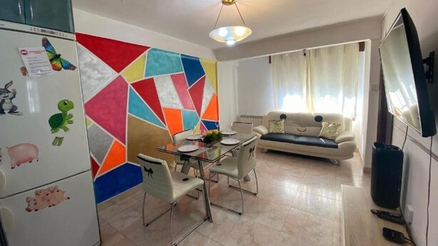 Sala de estar com sofá, mesa de jantar com cadeiras, televisão e frigorífico com desenhos de crianças.