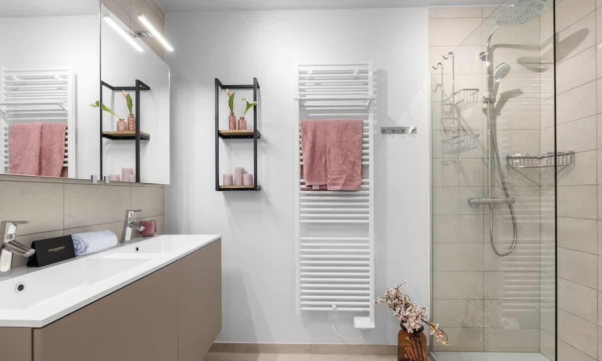 Un bany modern amb un mirall gran, un lavabo blanc, una cabina de dutxa de vidre i dues barraques per a tovalloles amb tovalloles roses.