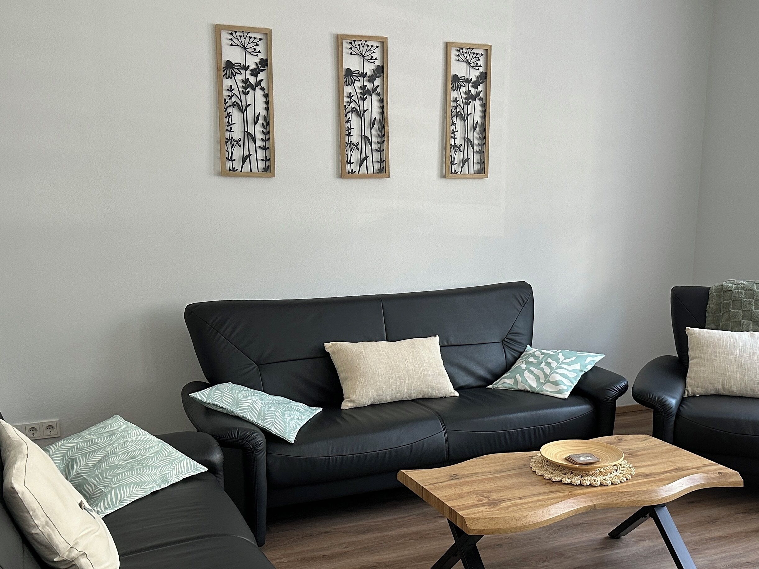 En moderne stue med en sort lædersofa, et træsofabord og tre billeder af botaniske motiver på væggen.
