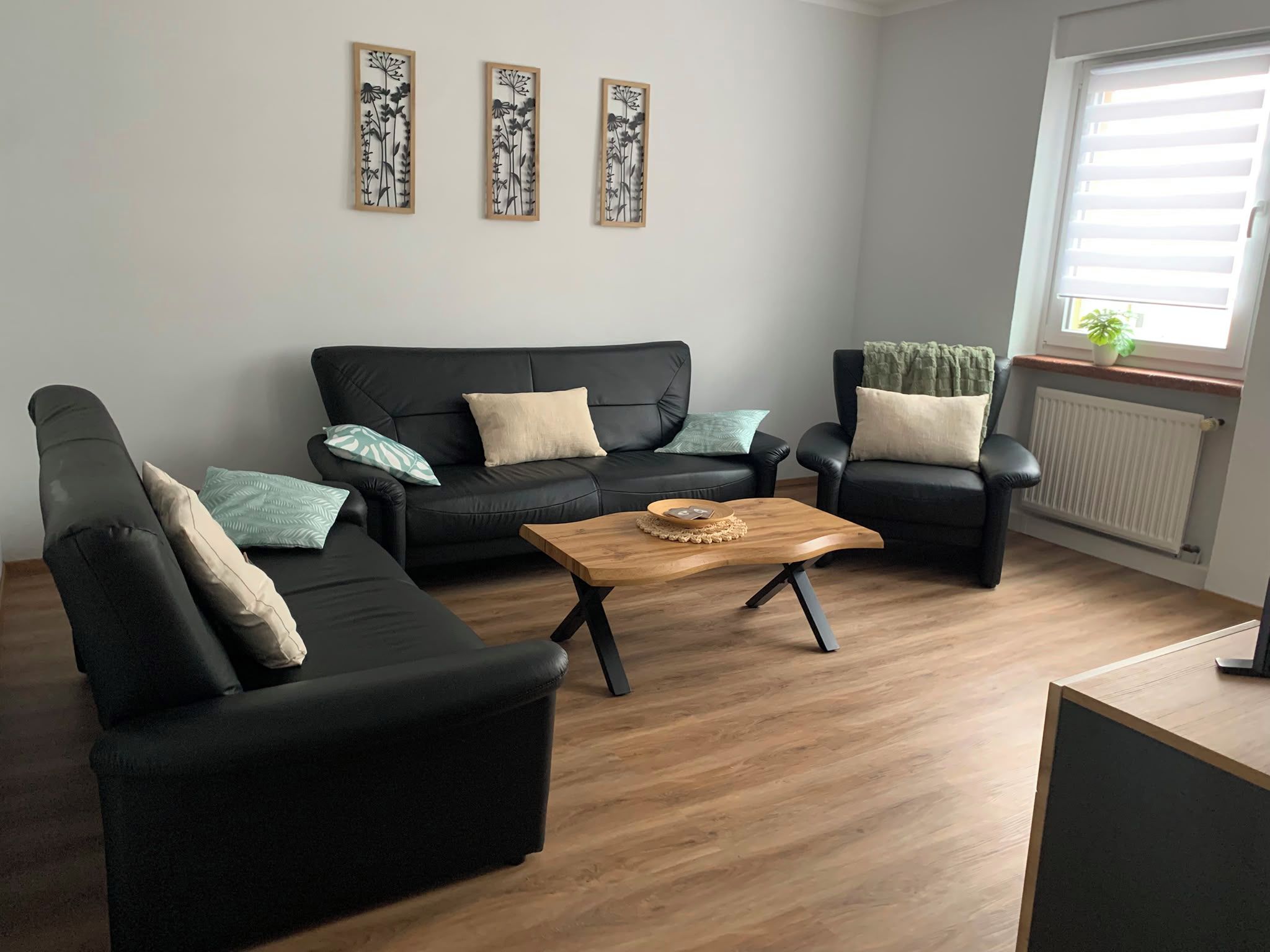 Ett vardagsrum med en svart soffa, ett träbord och en svart fåtölj. Ovanför soffan hänger tre dekorationer på väggen.