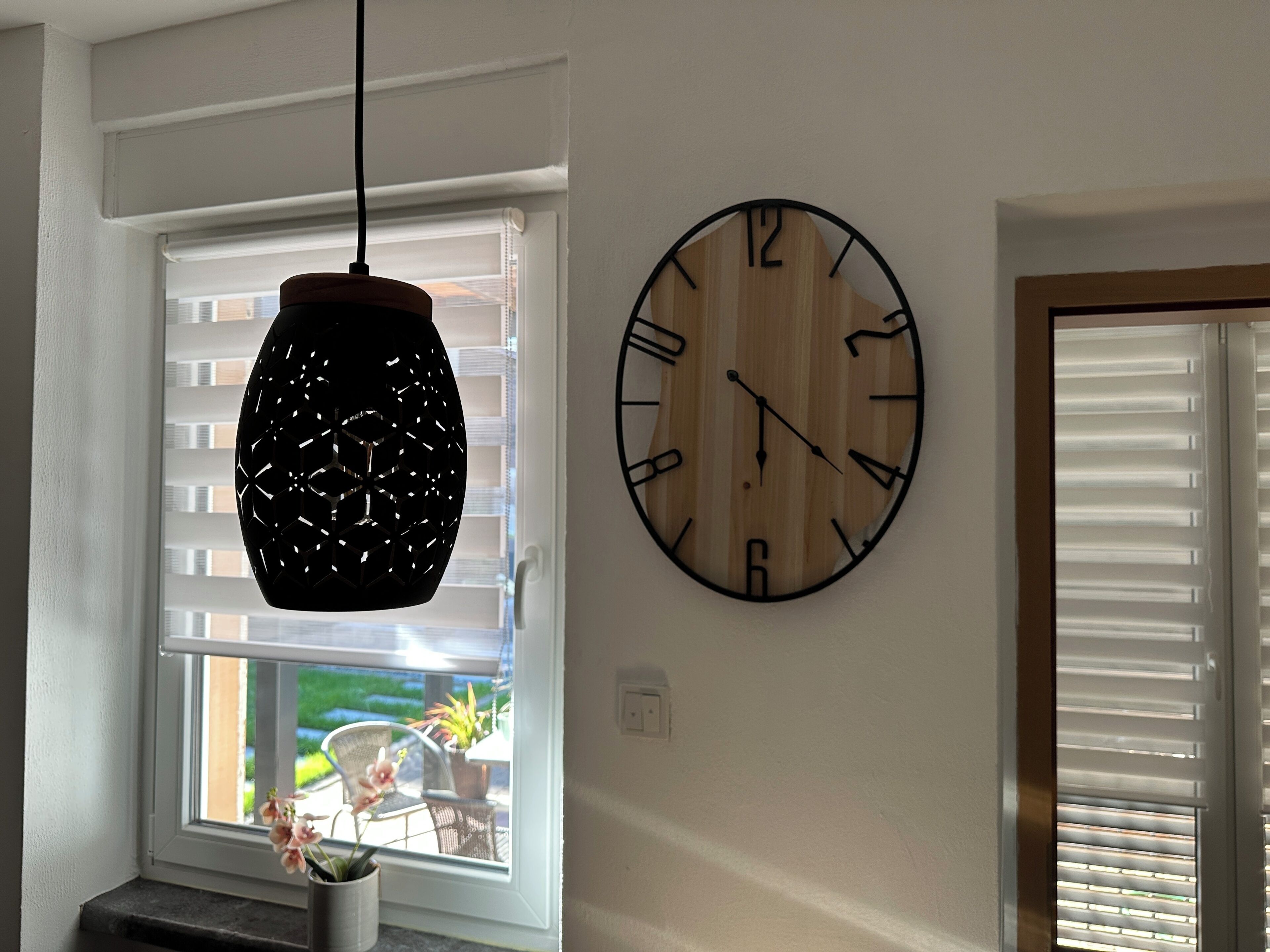 A room with a wooden wall clock, a hanging pendant light, and a window with blinds.
