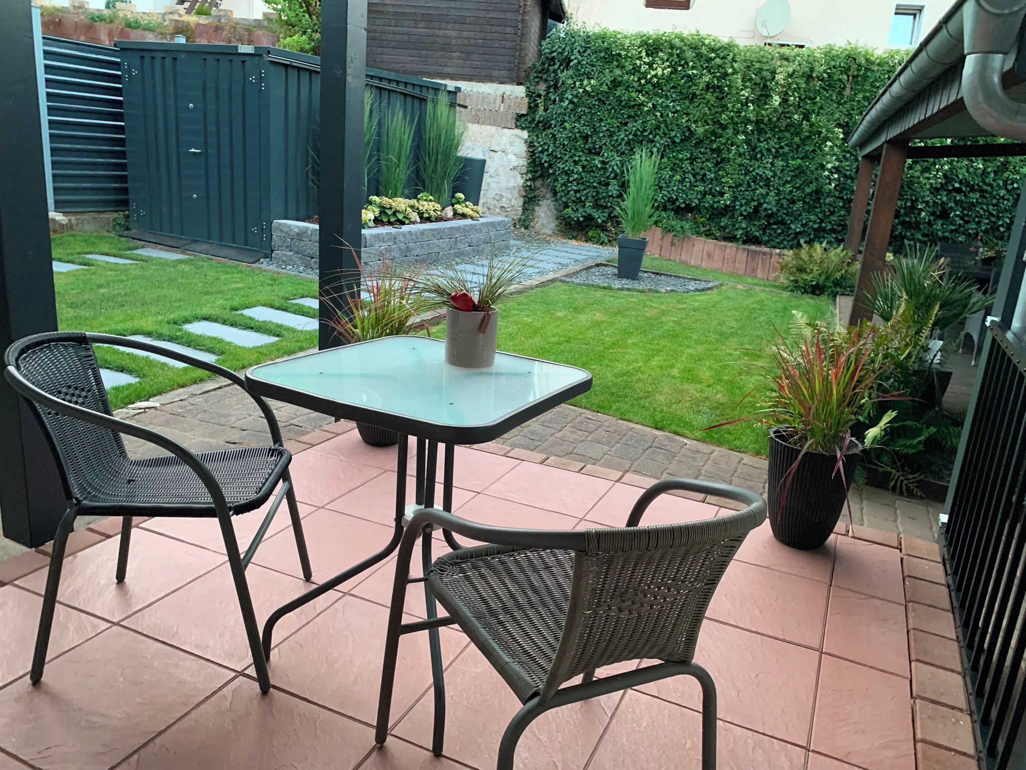 En udendørs patio med et bord og to stole, omgivet af en have med en hæk og krukker med planter.
