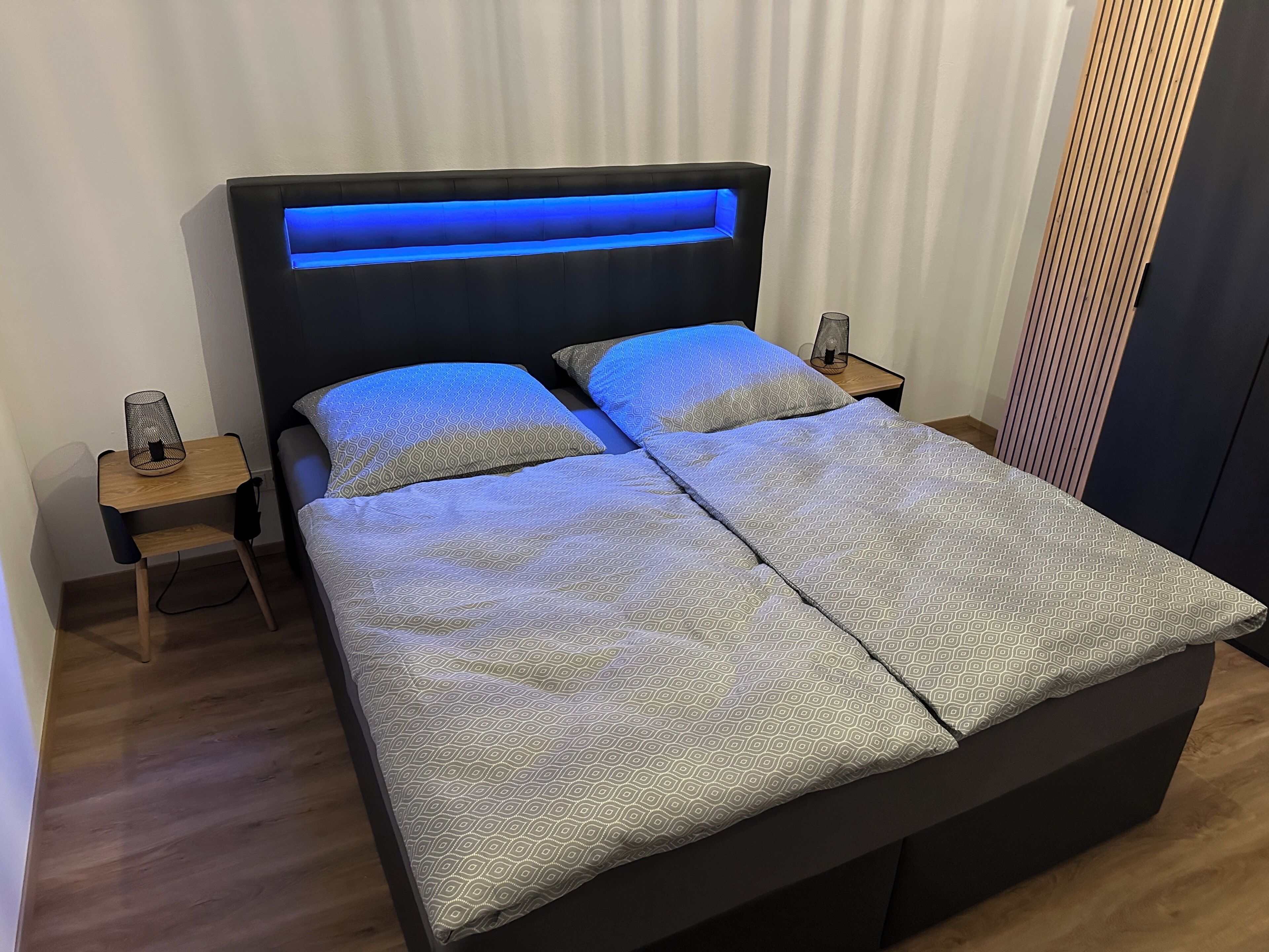 A double bed with a patterned bedspread, a headboard with blue lighting, two bedside tables with lamps, and a wooden floor.