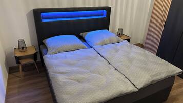 A double bed with a patterned bedspread, a headboard with blue lighting, two bedside tables with lamps, and a wooden floor.