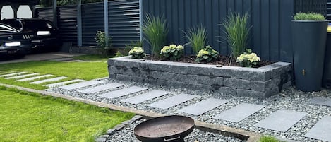 Un jardín con un camino de grava, un banco de metal y un parterre elevado con plantas.