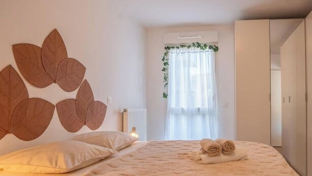 Ein Schlafzimmer mit Bett, Kissen, einer flauschigen Decke, einer Wanddekoration, einem Heizkörper, einem Fenster mit Vorhängen und einem Kleiderschrank.