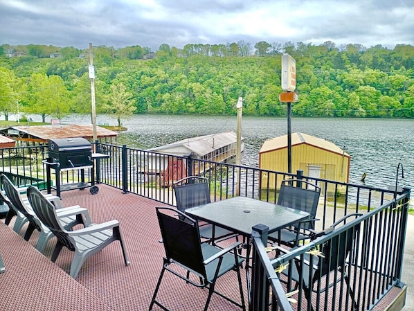 A deck with outdoor seating and a grill, overlooking a lake with a dock and a small shed.