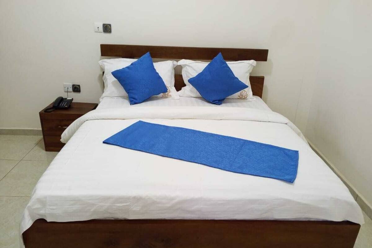 A bed with white bedding and two blue pillows, a wooden headboard, a bedside table with a phone, and a blue towel on the bed.