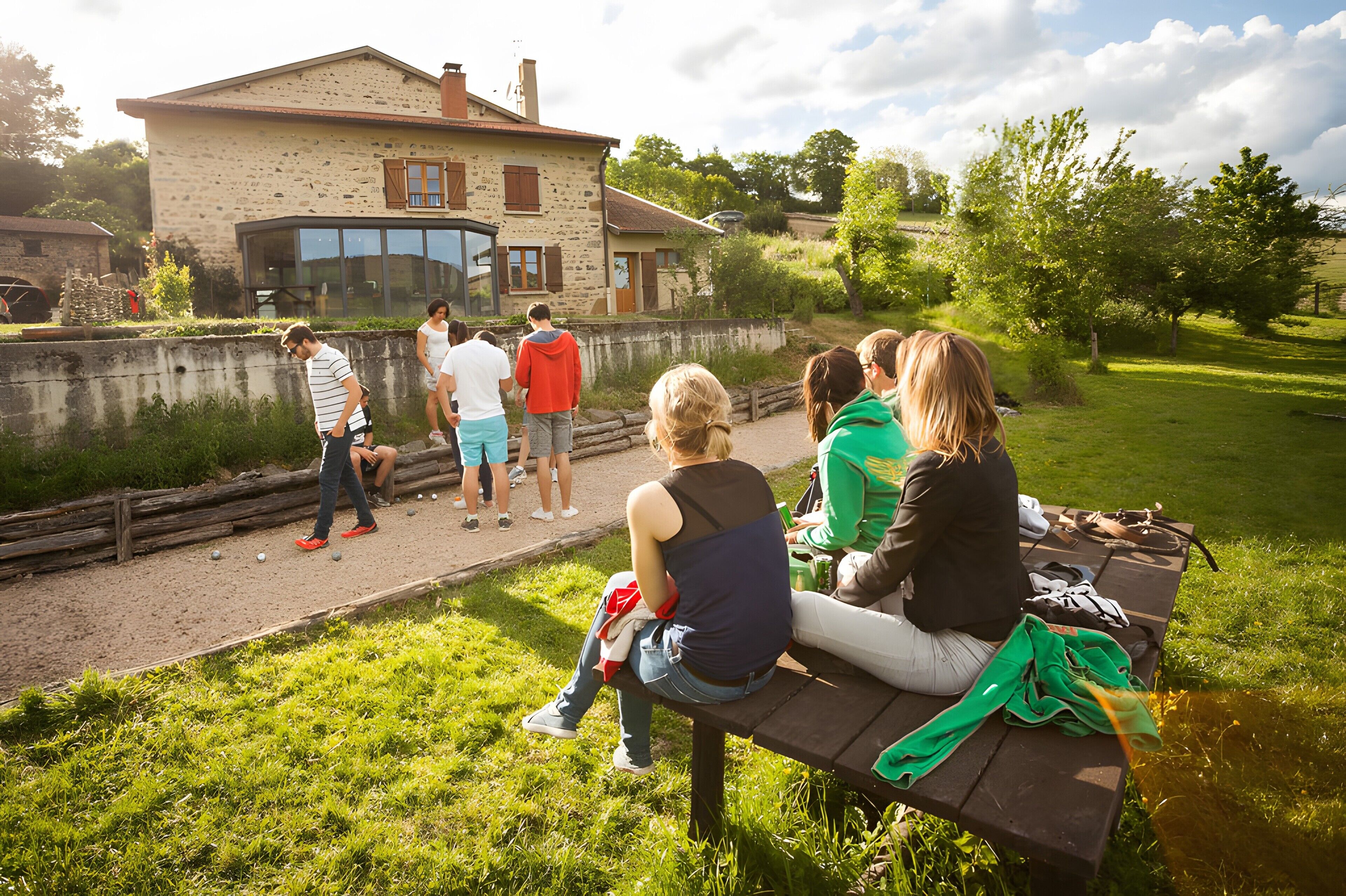 Eine Gruppe Menschen sitzt auf einer Bank im Freien, im Hintergrund ein Steinhaus und eine grüne Wiese.