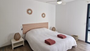 Ein Schlafzimmer mit einem Bett, einem Deckenventilator, zwei Wandspiegeln, einem Nachttisch, einem Korbwäscher und einem Fenster.