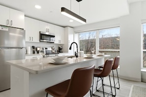 A modern kitchen with white cabinetry, stainless steel appliances, a central island with bar stools, and a large window offering a view of the outdoors. - Central & Comfortable Two Bedroom Two Bathroom (Cleveland)