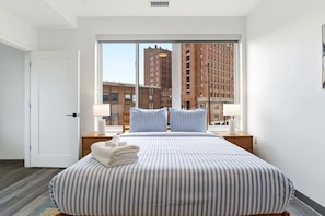 A hotel room with a large bed, two bedside lamps, a view of tall buildings, and a door to the left. - Relaxing & Central Two Bedroom Two Bathroom (Cleveland)