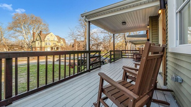 A wooden deck with a covered porch, a rocking chair, and a view of a residential area with houses. - Cozy & Spacious Four Bedroom (Cleveland)