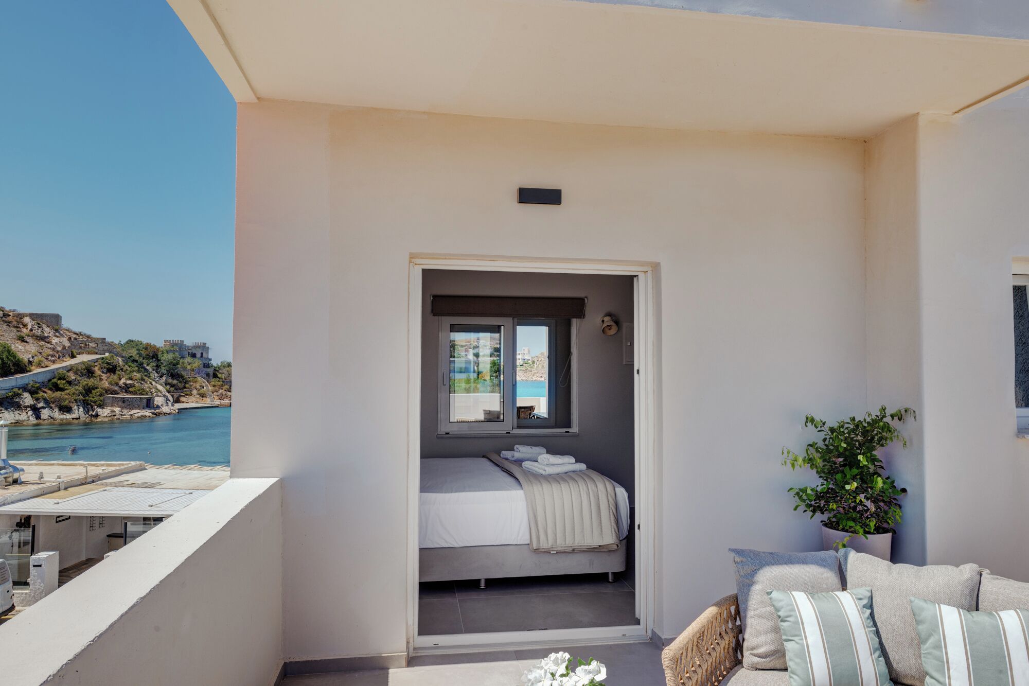A balcony with a bed, a potted plant, and a view of the sea and buildings.