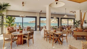 Un vaste espace dédié à la salle à manger, avec de nombreuses tables et chaises, de grandes fenêtres offrant une vue sur la mer et un plafond en bois.