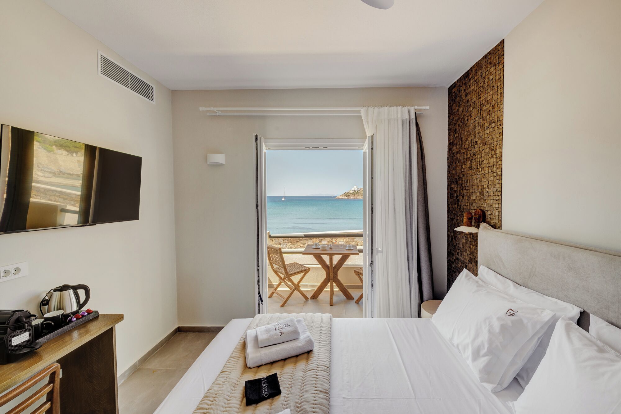 A hotel room with a bed, a desk with a kettle, a TV, and a view of the sea through an open door.