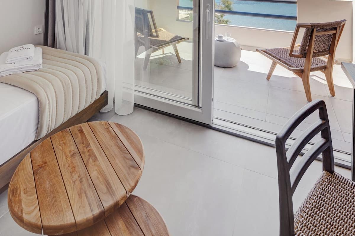 A modern hotel room with a bed, a wooden table, a chair, and a balcony with a view of the sea.