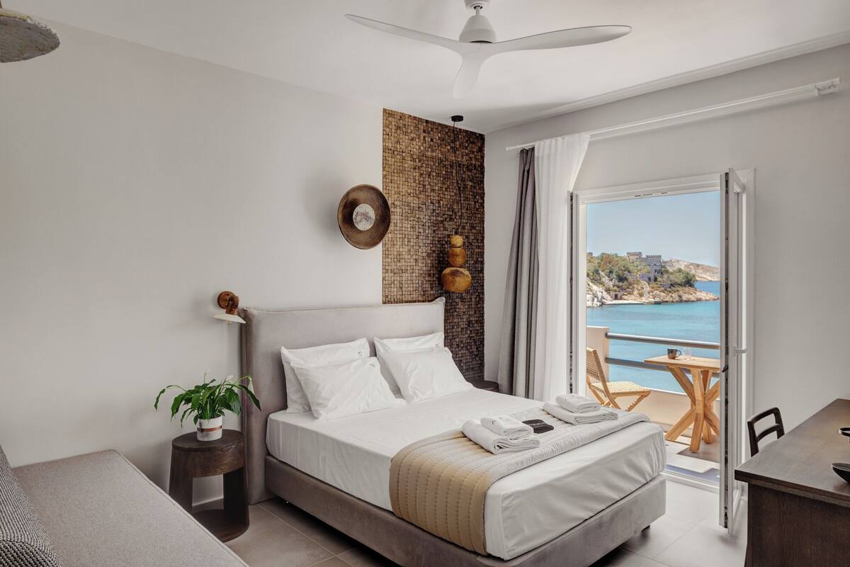 A bedroom with a bed, a potted plant, a wooden side table, a view of the sea, and a balcony with a table and chairs.