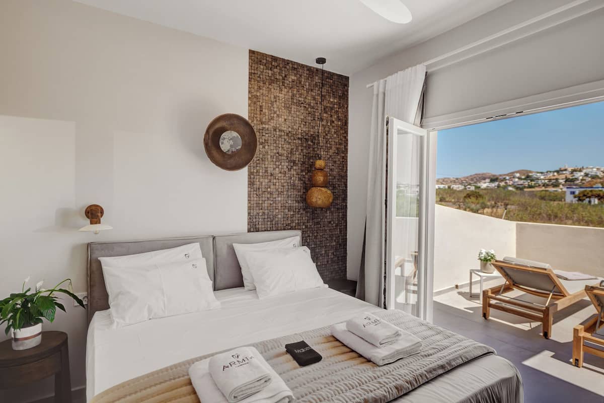 A bedroom with a bed, a bedside table with a plant, a wall-mounted clock, and a view of a balcony with a chair and a table.