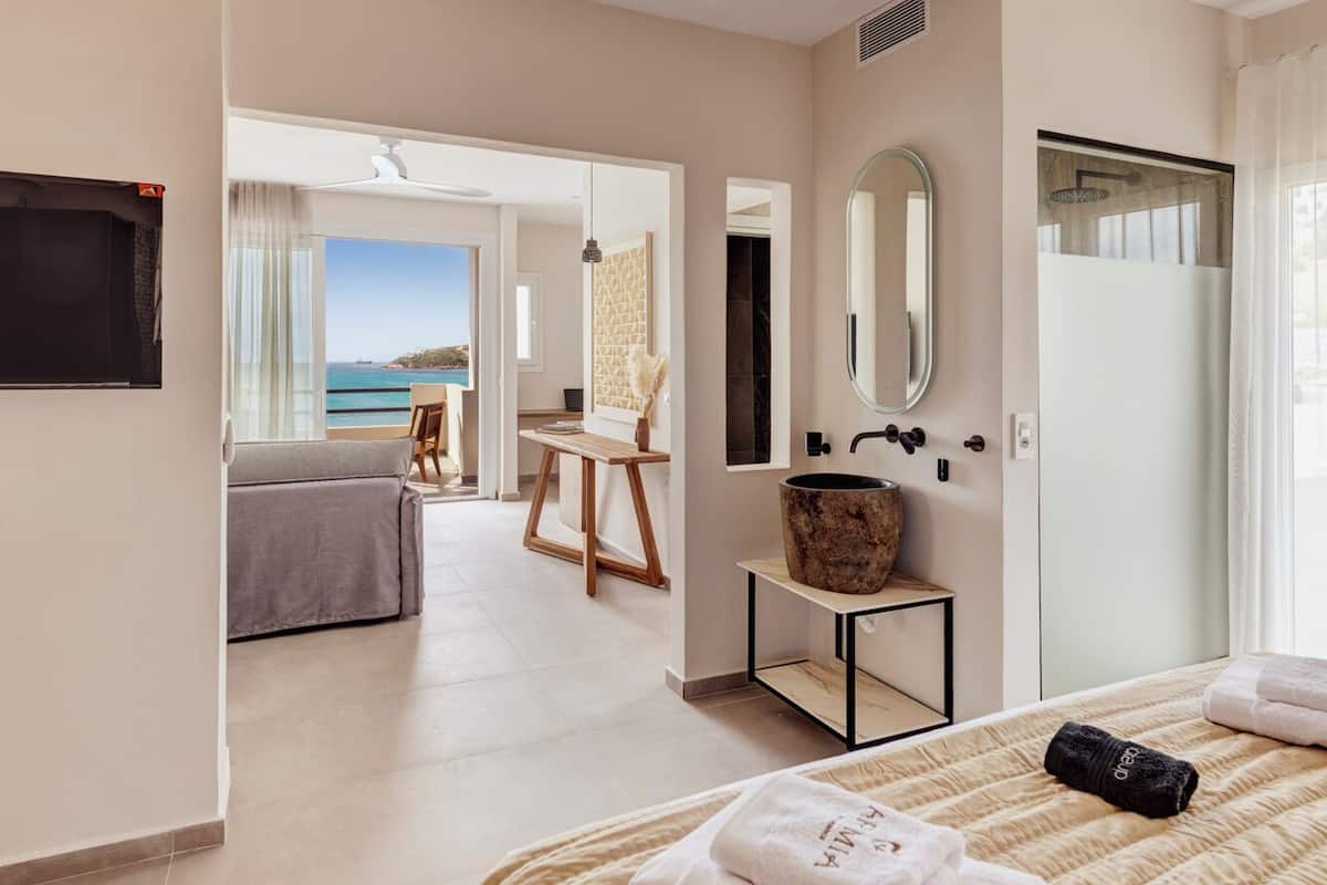 A modern hotel room with a large bed, a ceiling fan, a bathroom with a shower, and a view of the sea.