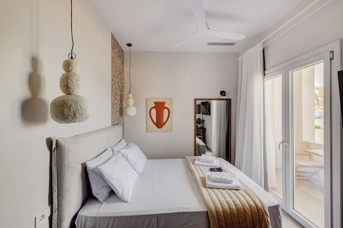 A bedroom with a bed, a ceiling fan, a wall-mounted light fixture, a mirror, and a window with a view.