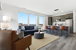 Eine moderne Wohnung mit Wohnzimmer, Sofa, Couchtisch, Küche mit Barhocker und großen Fenstern.