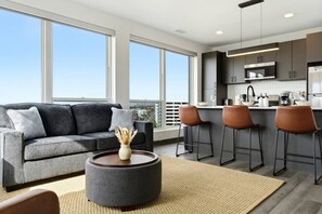 Ein modernes Wohnzimmer mit grauem Sofa, einem runden Couchtisch und Blick auf die Stadt durch große Fenster.