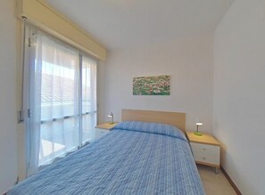 A bedroom with a bed, two bedside tables, a window with curtains, and a wall-mounted picture. - Seaside Escape for Four Guests (Lignano Sabbiadoro)