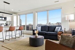 A modern living room with a sofa, coffee table, and chairs, large windows with a city view, and a kitchen area. - Charming & Airy Two Bedroom Two Bathroom (Cleveland)