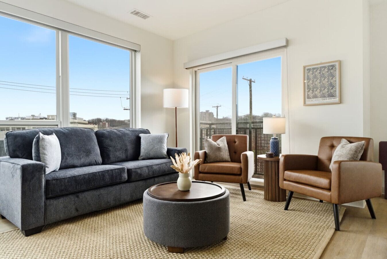 A modern living room with a grey sofa, two armchairs, a round coffee table, and large windows offering a city view.