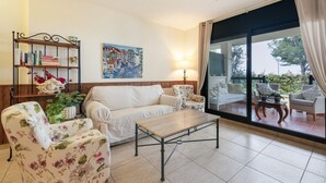 Un salón con sofá, sillones, una mesa de centro, una estantería, un cuadro y vistas a través de un ventanal.