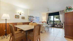 Un comedor con sillas de mimbre, una mesa y una lámpara. Se observa una estantería con fotografías enmarcadas y un ventilador de techo.