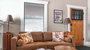 A living room with a brown leather sofa, two side tables with lamps, a wooden door with a black curtain, and a poster on the wall. - Central Tucson Summer Retreat | Near UofA (Tucson)