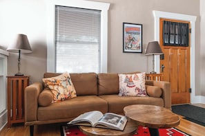 Ein Wohnzimmer mit einem braunen Ledersofa, zwei Beistelltischen mit Lampen, einer Holztür mit schwarzem Vorhang und einem Poster an der Wand.