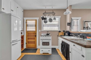 Eine kompakte Küche mit einem weißen Kühlschrank, Herd, Backofen und einer Mikrowelle. Ein Fenster mit Jalousien, ein Spülbecken und eine Arbeitsplatte mit Geschirrständer sind vorhanden.