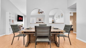 Uma sala de jantar com uma mesa de madeira, quatro cadeiras, um ventilador de teto e uma TV na parede.