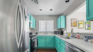 A modern kitchen with teal cabinets, stainless steel appliances, and a window with blinds.